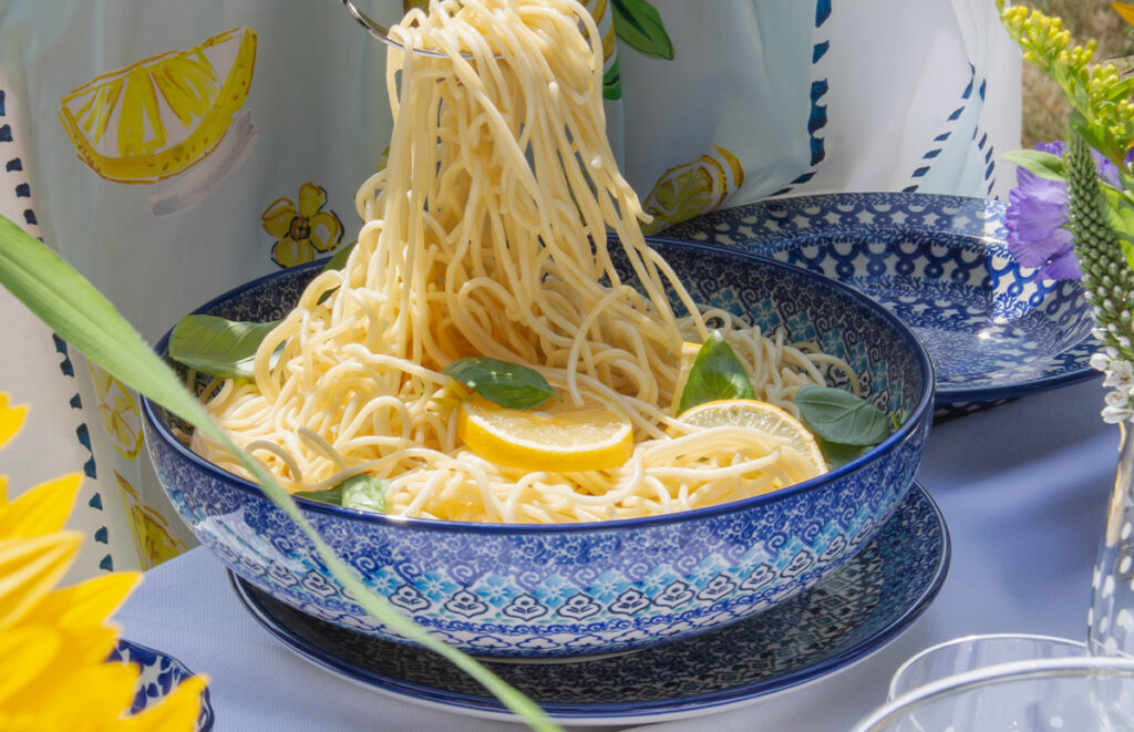 Close-up of a Bunzlau Castle serving bowl filled with spaghetti, garnished with fresh basil and lemon slices, set on a beautifully styled outdoor dining table.
