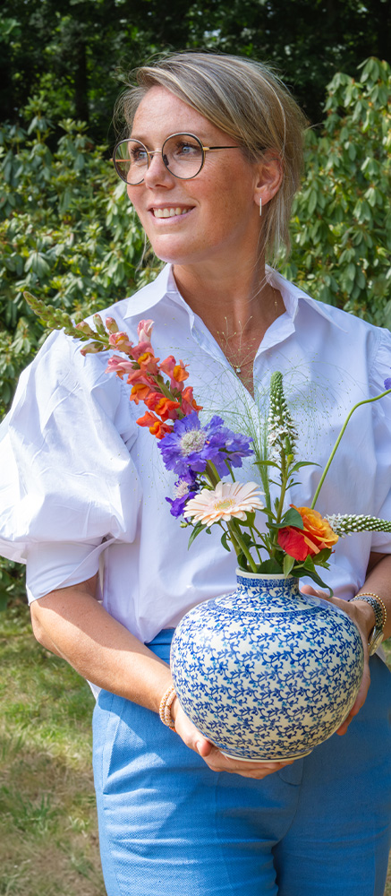 A woman is holding a new Bunzlau Castle vase filled with a fresh bouquet of flowers, standing in a beautiful lush green outdoor setting.